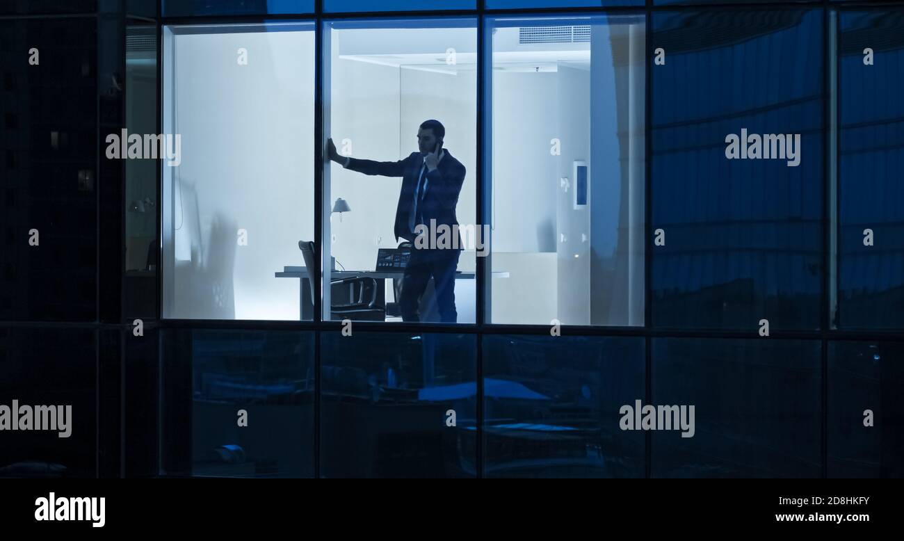 Aerial Shot: From Outside into Office Building with Businessman Using Mobile Phone and Standing by the Office Window. Beautiful Shot of The Financial Stock Photo