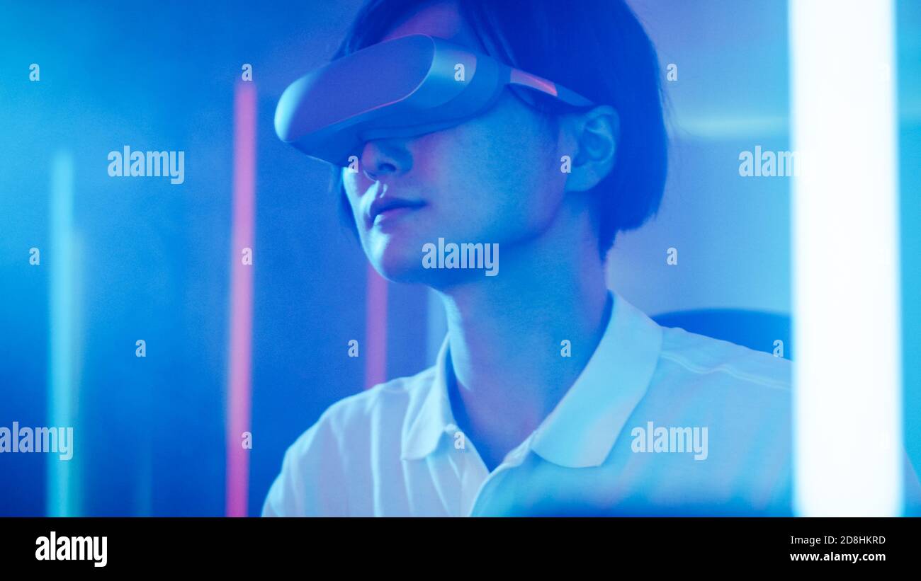 East Asian Pro Gamer Wearing Virtual Reality Headset Plays Online Video Game with Joysticks Controllers. Cool Retro Neon Colors in the Room. Stock Photo