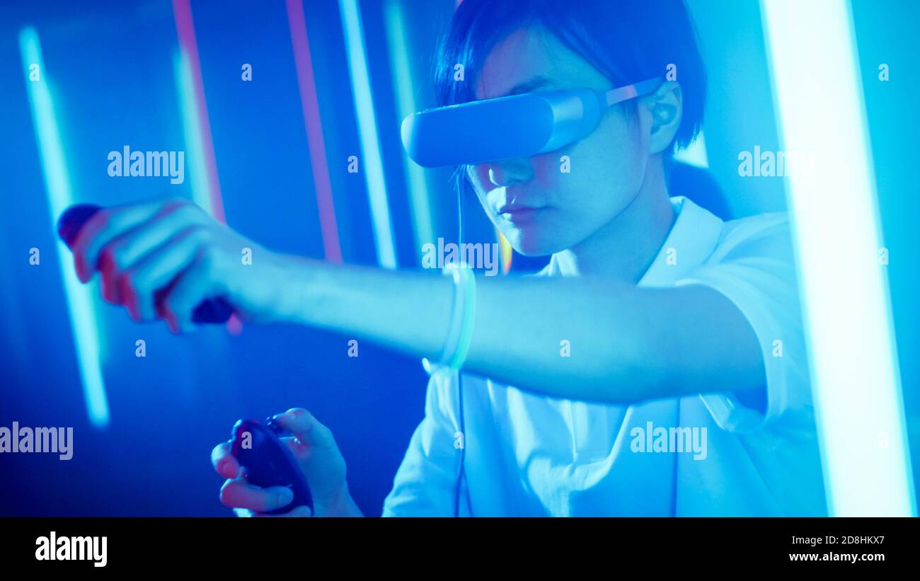 East Asian Pro Gamer Wearing Virtual Reality Headset Plays Online Video Game with Joysticks Controllers. Cool Retro Neon Colors in the Room. Stock Photo