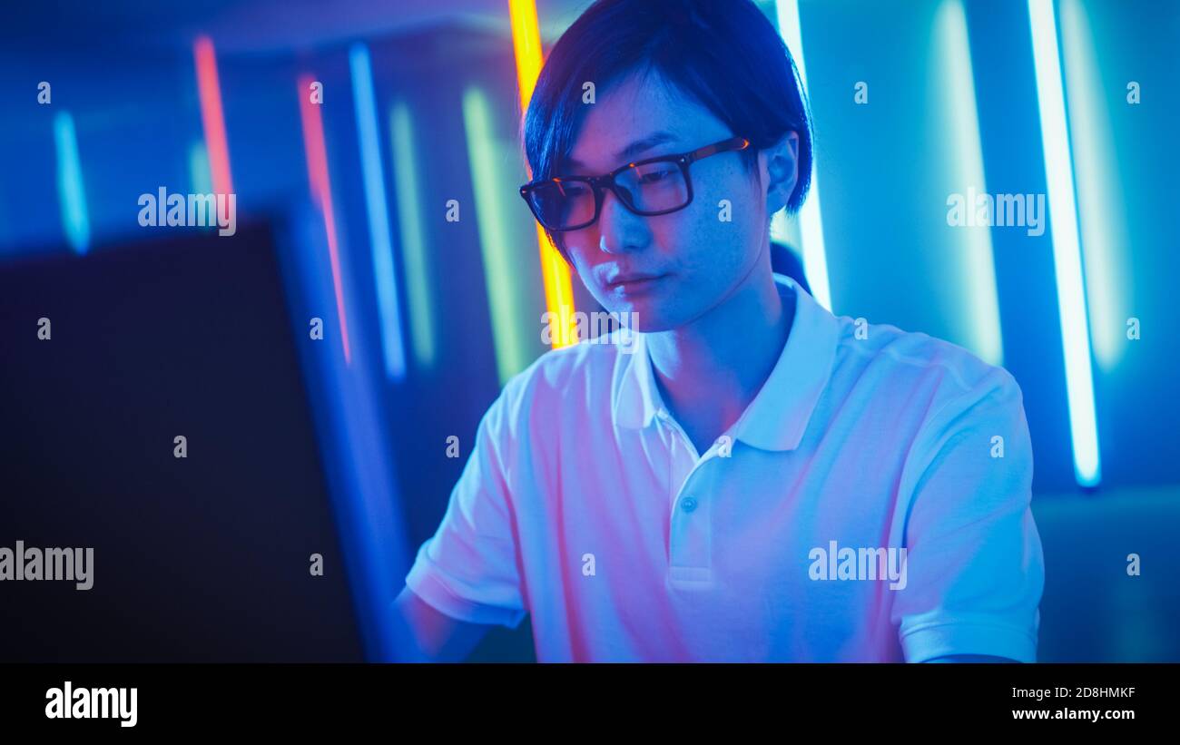 Handsome Young Guy Sits Before Personal Computer, Working, Surfing in Internet and Playing Online Games. Cool Neon Retro Lights Behind Him. Stock Photo