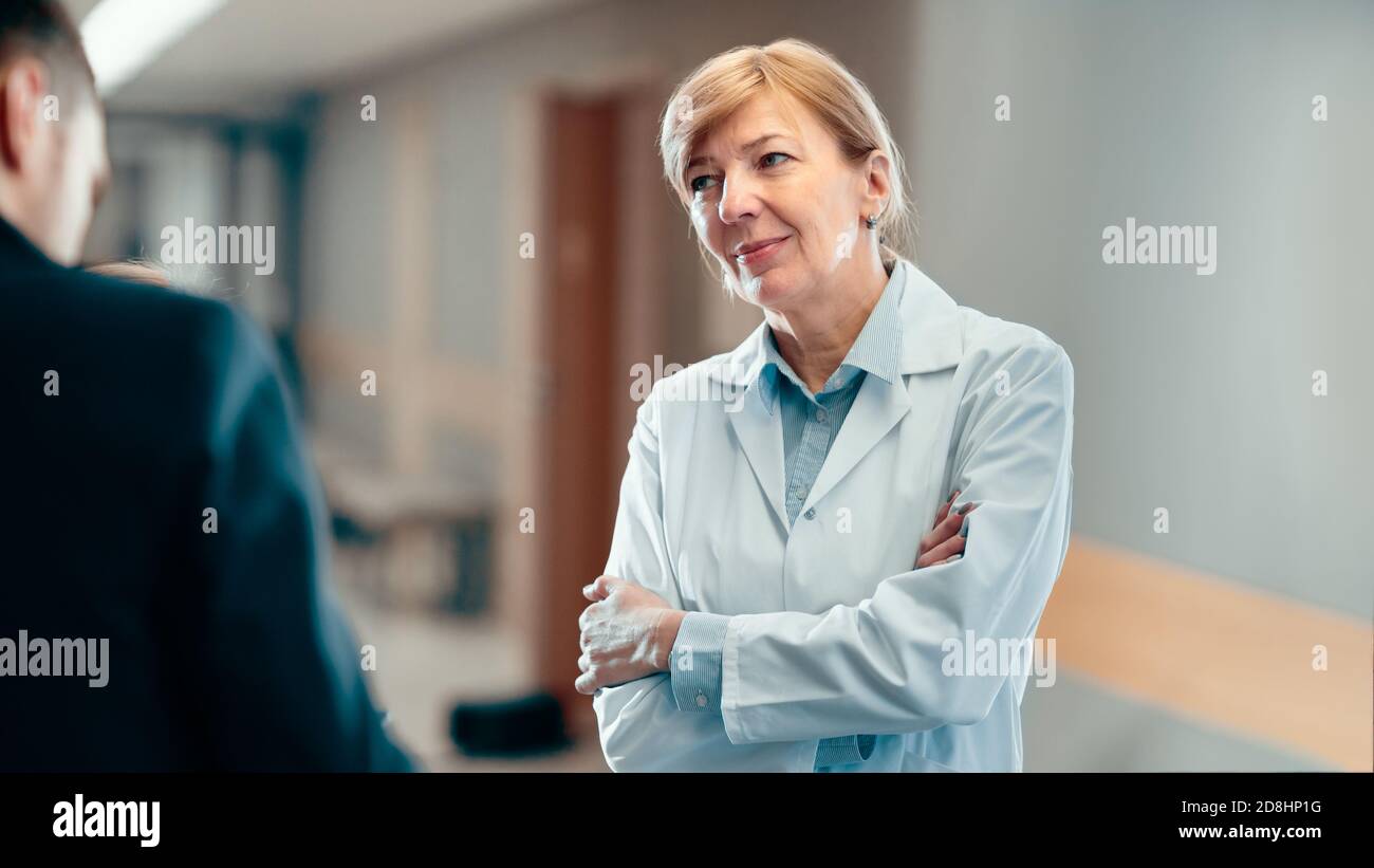 Portrait Shot of a Senior Female Doctor Standing in the Hospital Hallway and Having Professional Discussion. Modern Hospital with Patients and Medical Stock Photo