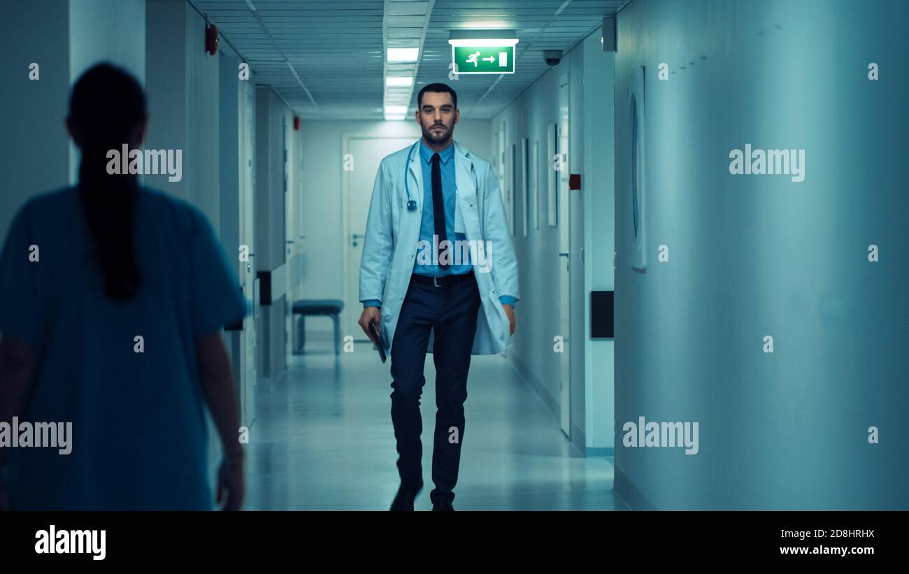 Determined Handsome Doctor Wearing White Coat with Stethoscope Walks Through Hospital Hallway. Modern Bright Clinic with Professional Staff. Stock Photo