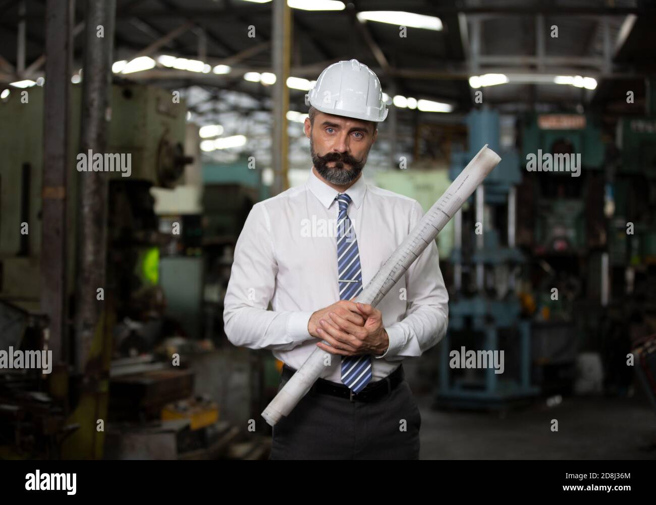 The bearded hipster director with engineer hat standing against machine in production in factory Stock Photo