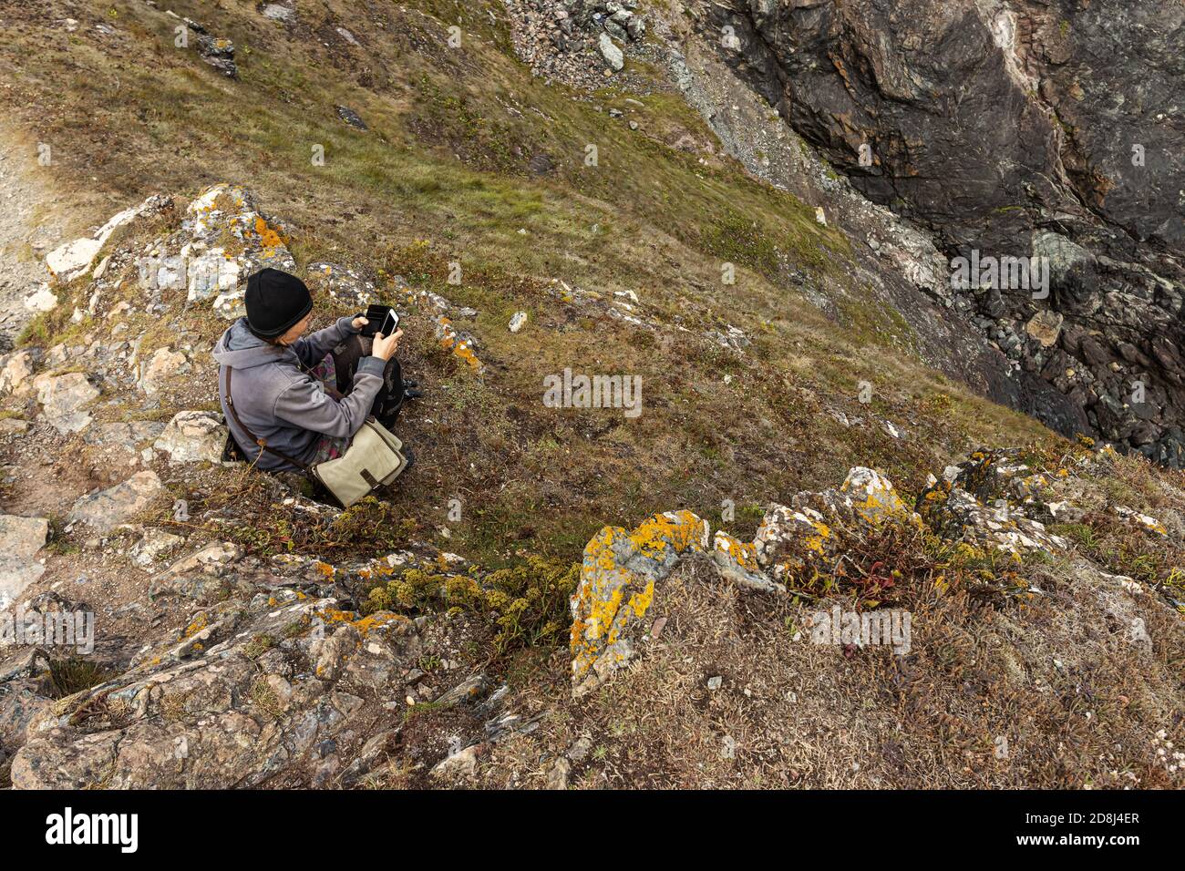 A female wrapped up in warm clothing watching sitting on a cliff and taking a photo with a phone Stock Photo