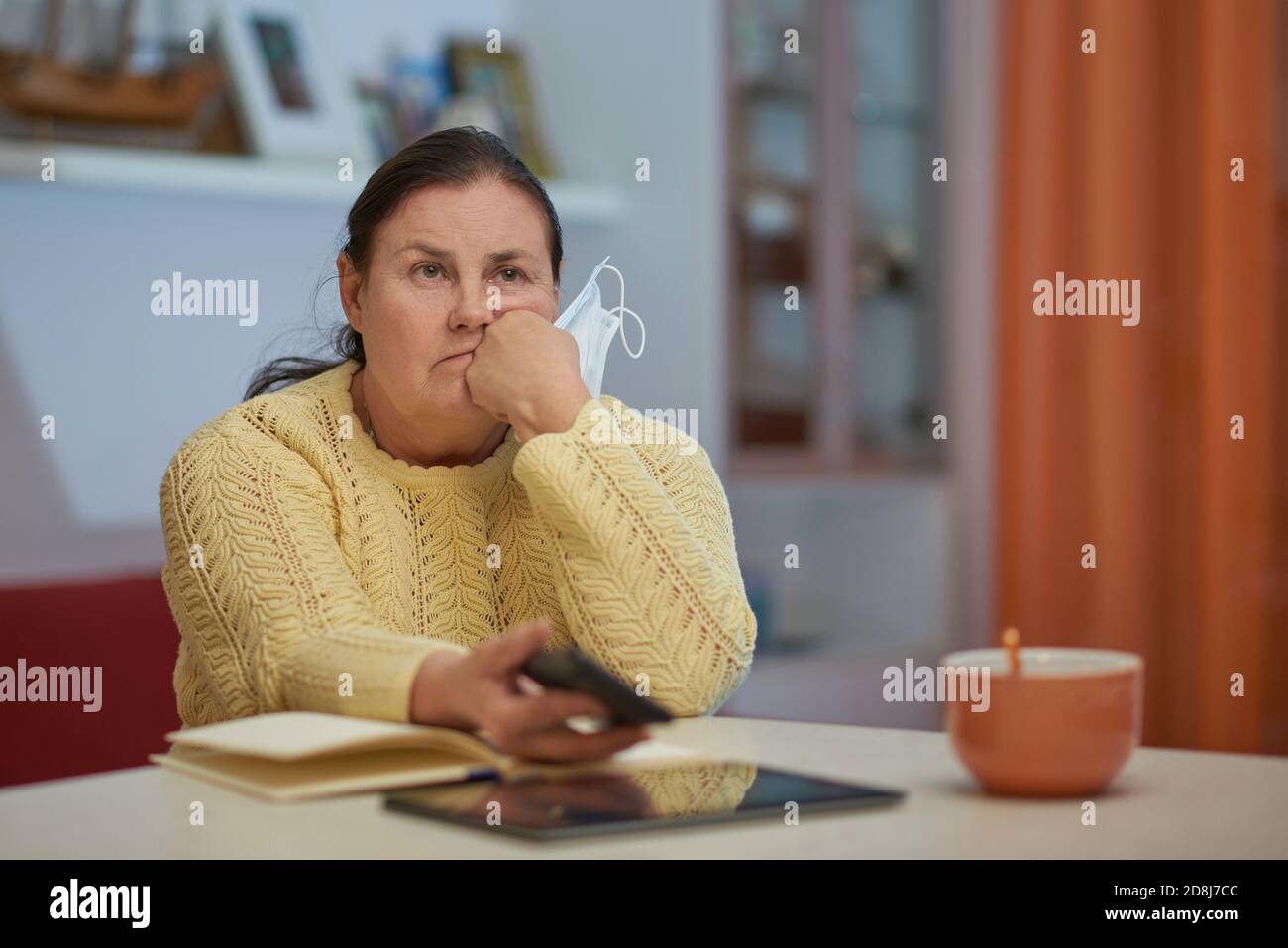 Sad senior woman in yellow sweater with mask and smart phone Stock Photo