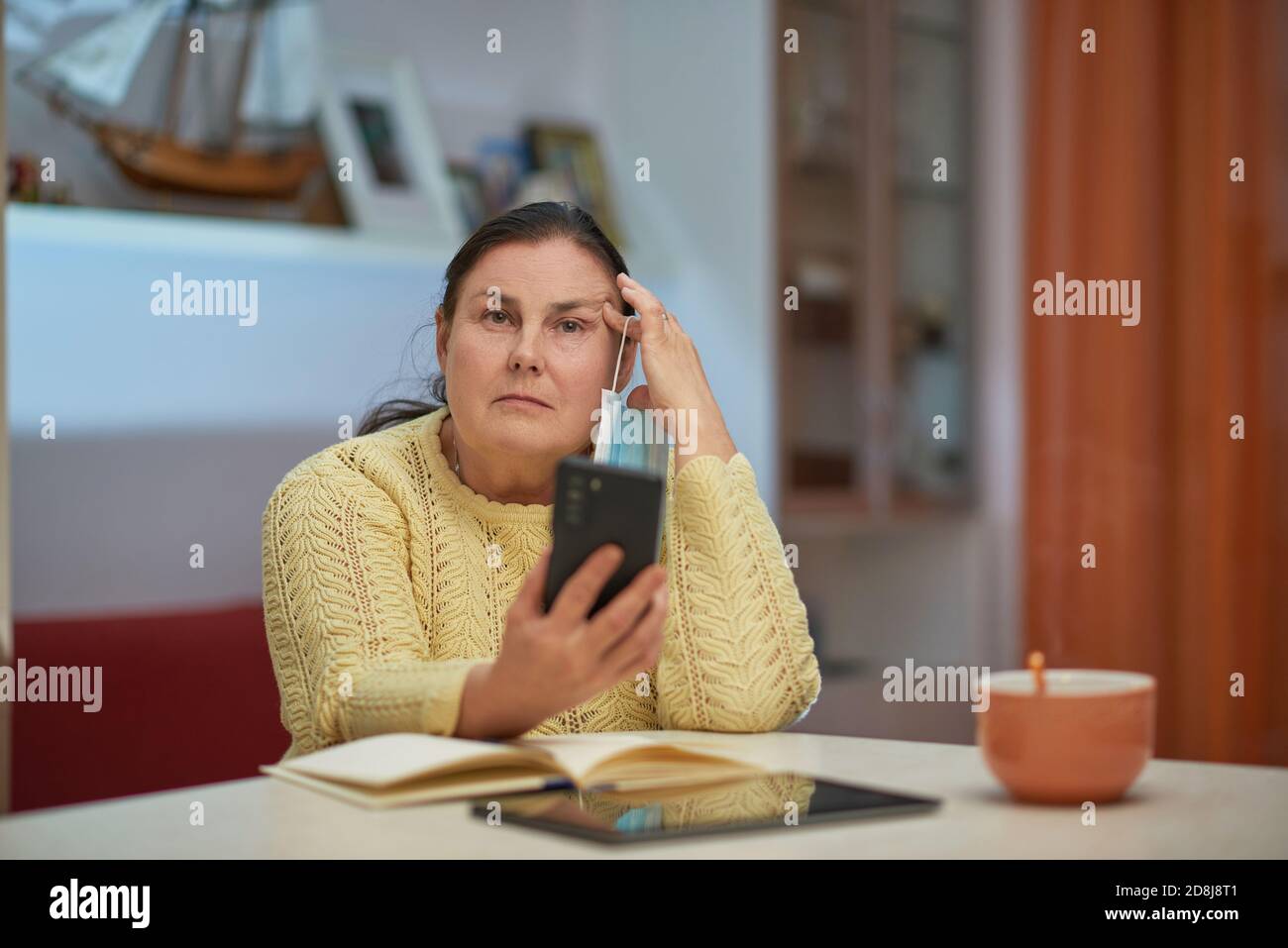 Sad senior woman in yellow sweater with smartphone and mask Stock Photo