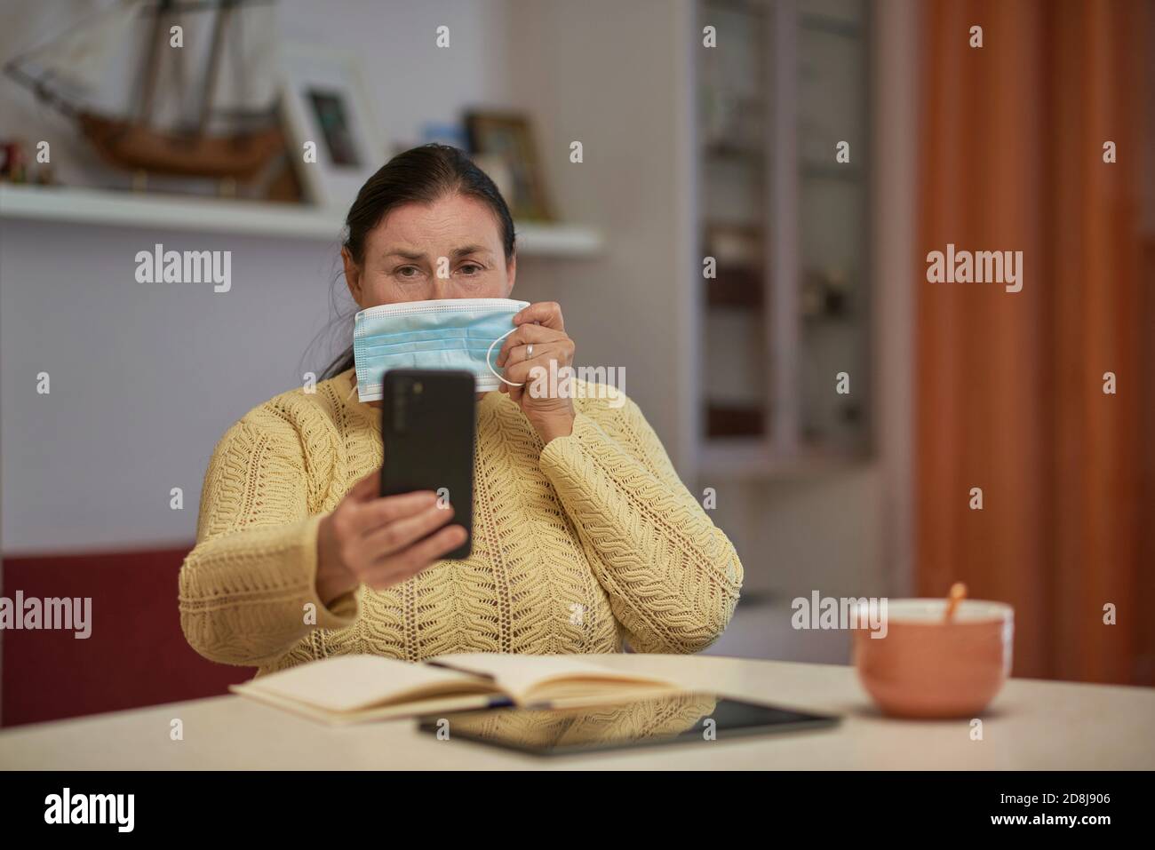 Senior woman in yellow sweater looking at smartphone with mask Stock Photo