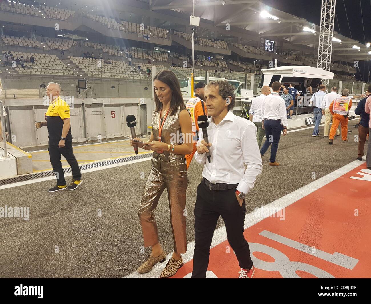 Famous ancient F1 driver Alain Prost walks by the Pit Line with a young female TV Presenter holding a microphone in her hands Stock Photo