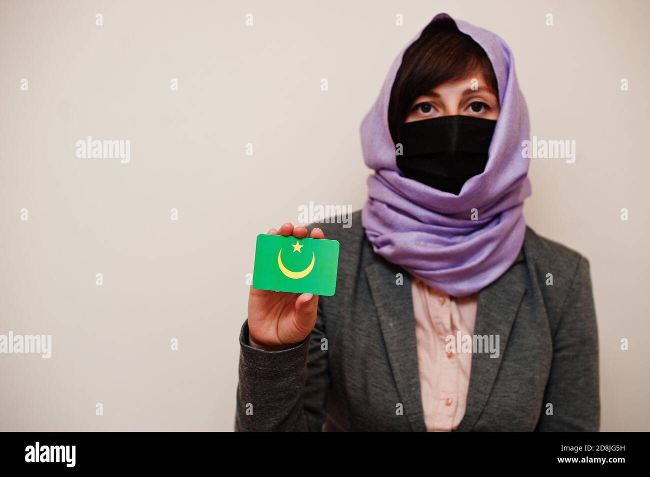 Portrait of young muslim woman wearing formal wear, protect face mask and hijab head scarf, hold Mauritania flag card against isolated background. Cor Stock Photo