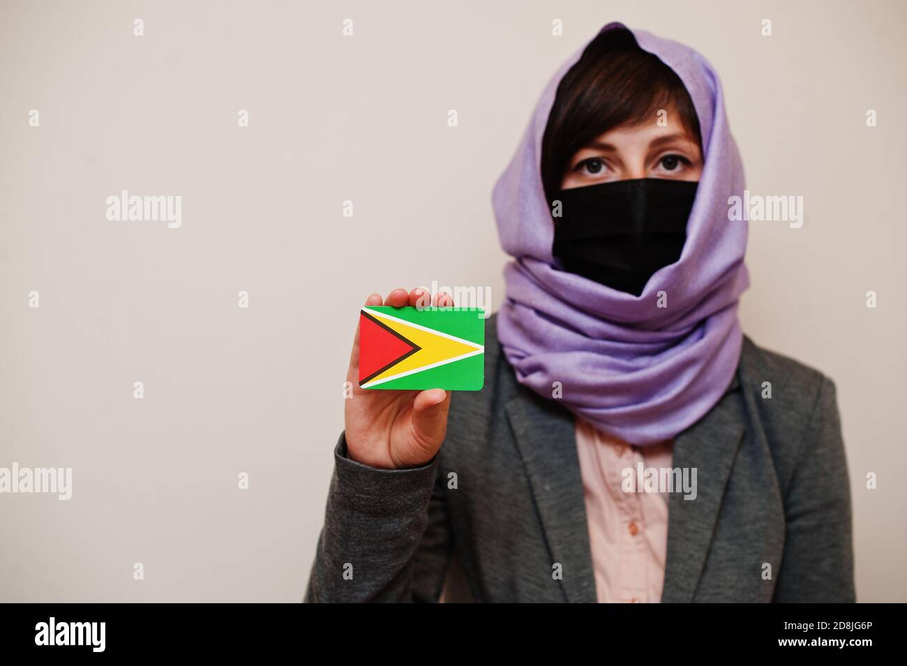Portrait of young muslim woman wearing formal wear, protect face mask and hijab head scarf, hold Guyana flag card against isolated background. Coronav Stock Photo