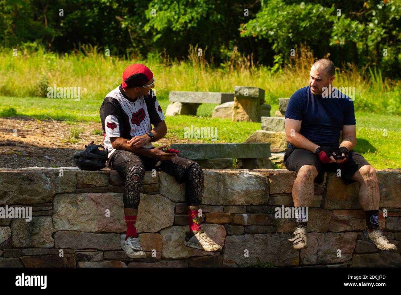 Maryland, USA 06/27/2020: Two men are resting on a stone wall in a forest and talking to eachoter. They wear cycling clothes and they have just finish Stock Photo