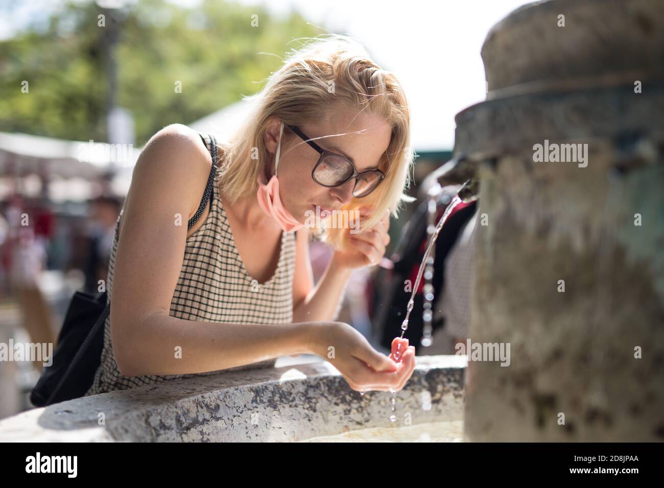 Thirsty young casual cucasian woman wearing medical face mask drinking water from public city fountain on a hot summer day. New social norms during Stock Photo
