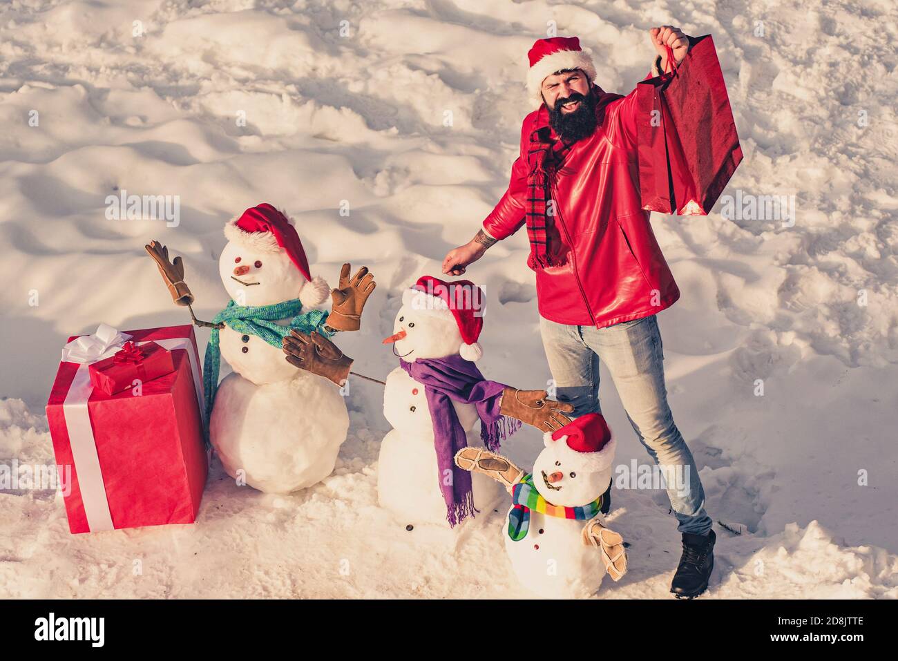 Excited man with funny snowman in stylish hat and scarf on snowy field. Happy winter snowman family with gift. Mother snow-woman, father snow-man and Stock Photo