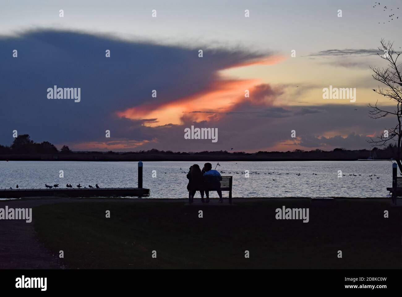 A couple sit on a bench watching the sunset at Oulton Broad near Lowestoft in The Broads National Park.  The clouds create a dramatic pattern. Stock Photo