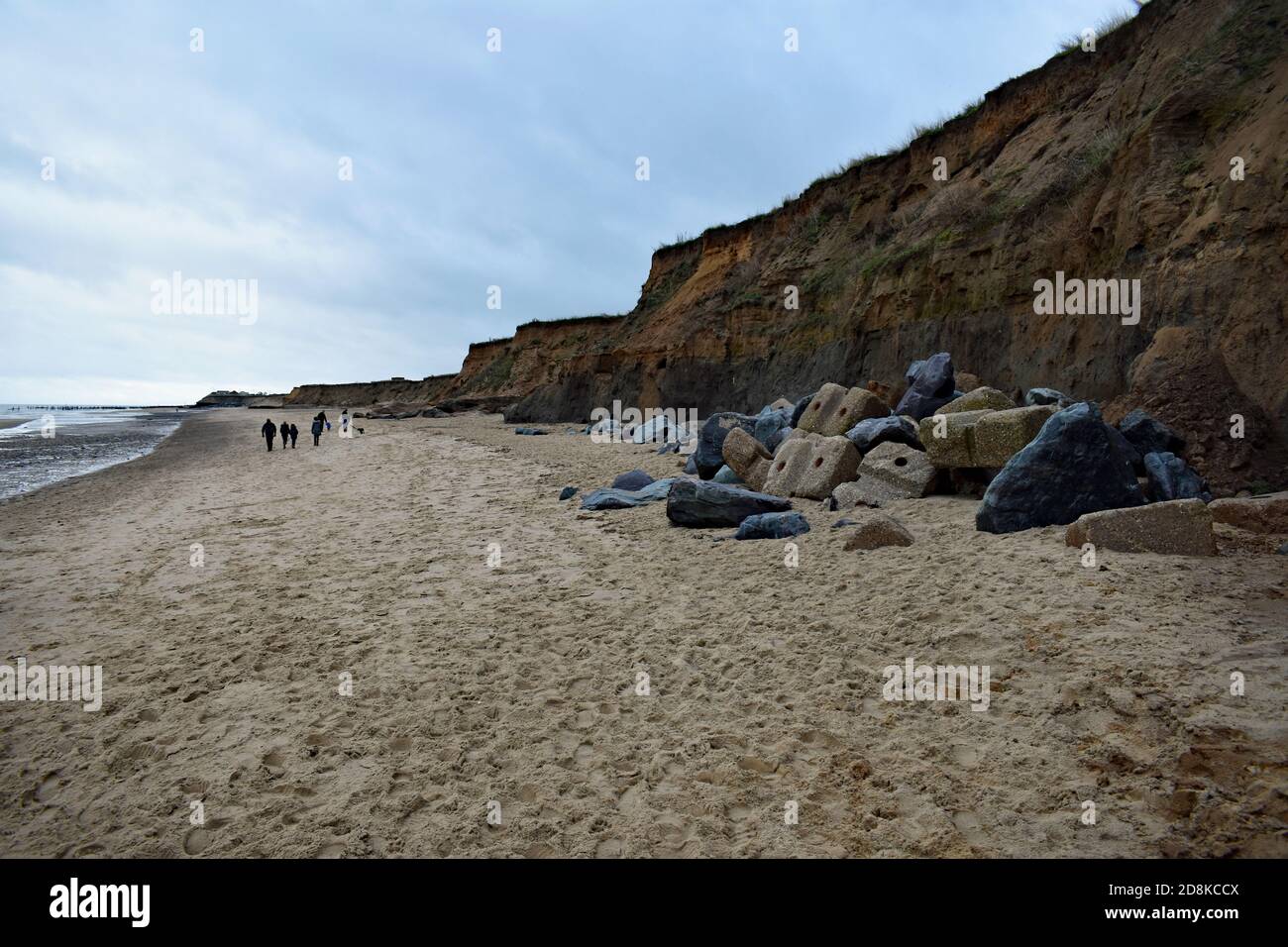A family walk along Happisburgh Beach next to the sea cliffs that are displaying costal erosion in Norfolk, England. Large rocks stand along the cliff. Stock Photo