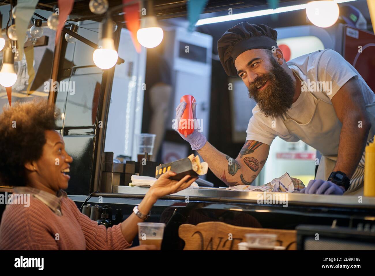 young beardy caucasian employee in fast food adding a ketchup in a sandwich to a female afro-american customer, joking, amusing Stock Photo