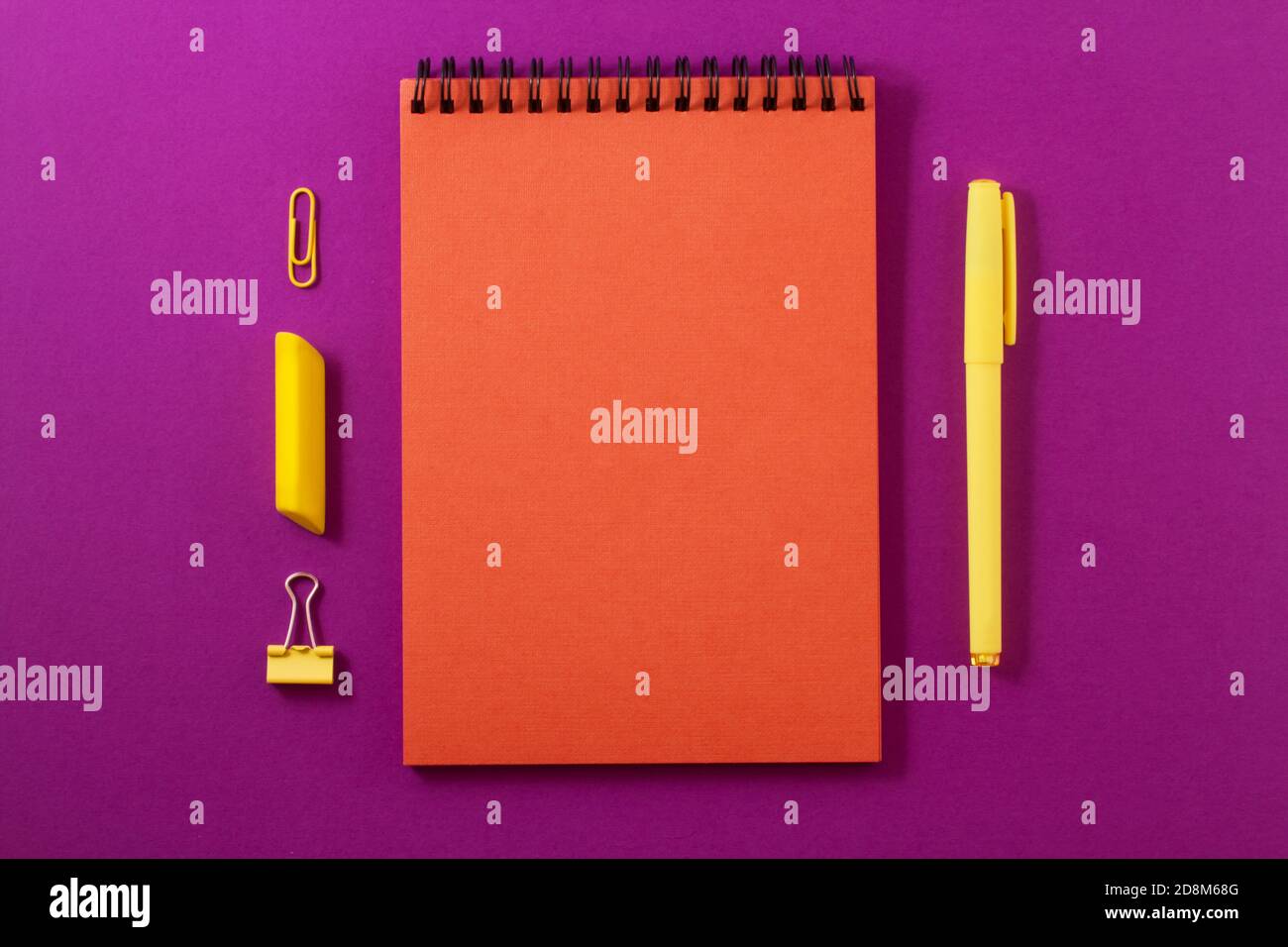 An orange notebook on a bright purple background with yellow office supplies on it, a pen and an eraser and a clamp and a clip. Stock Photo