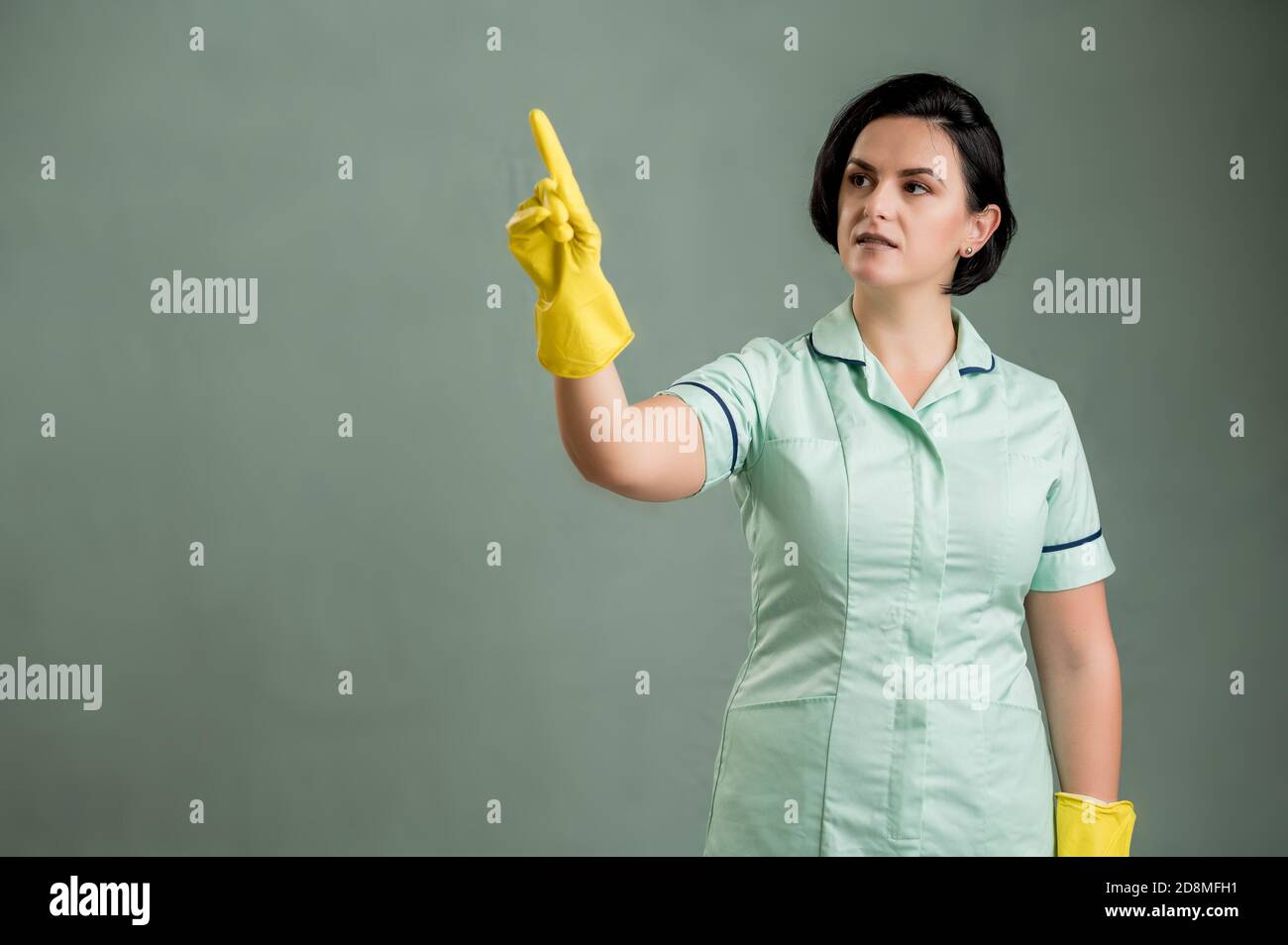 Young cleaning woman wearing a green shirt and yellow gloves, presses a virtual button isolated on green background Stock Photo