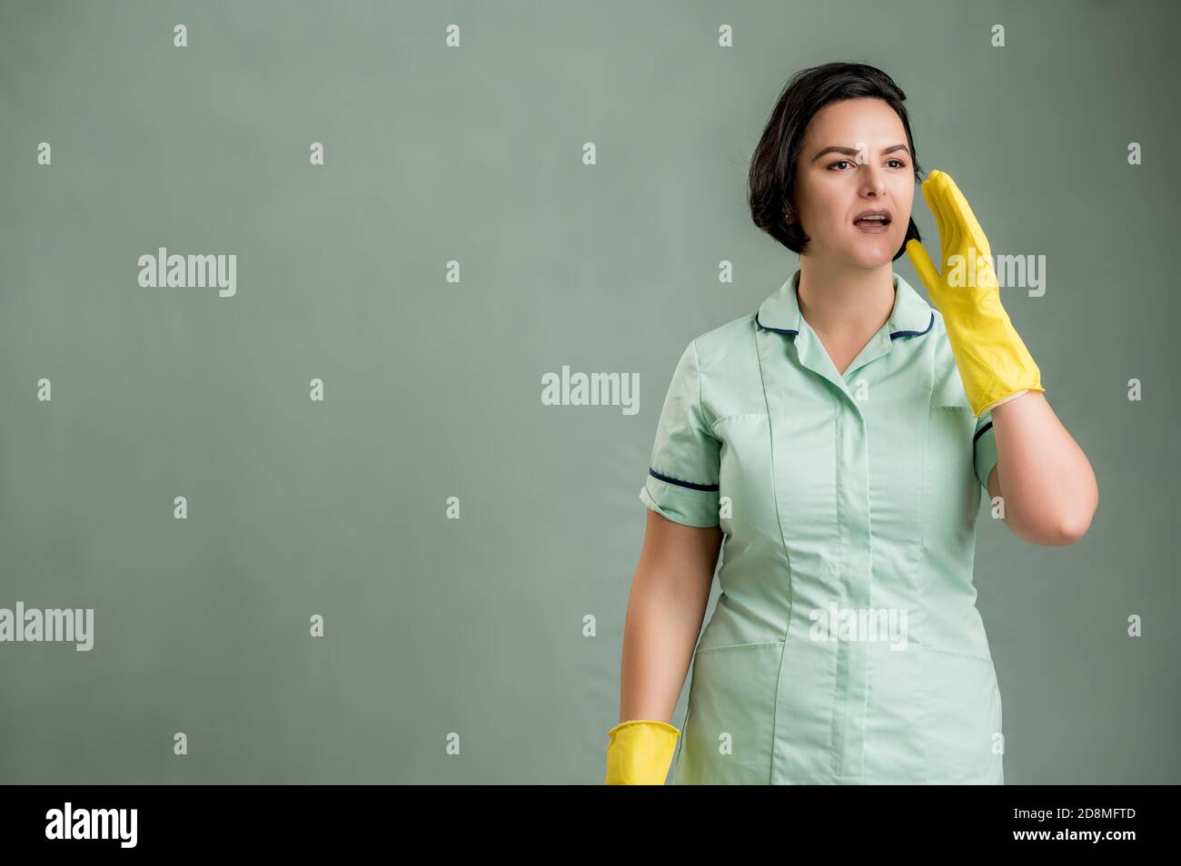Young cleaning woman wearing a green shirt and yellow gloves, shouting with her hand to her mouth isolated on green background Stock Photo