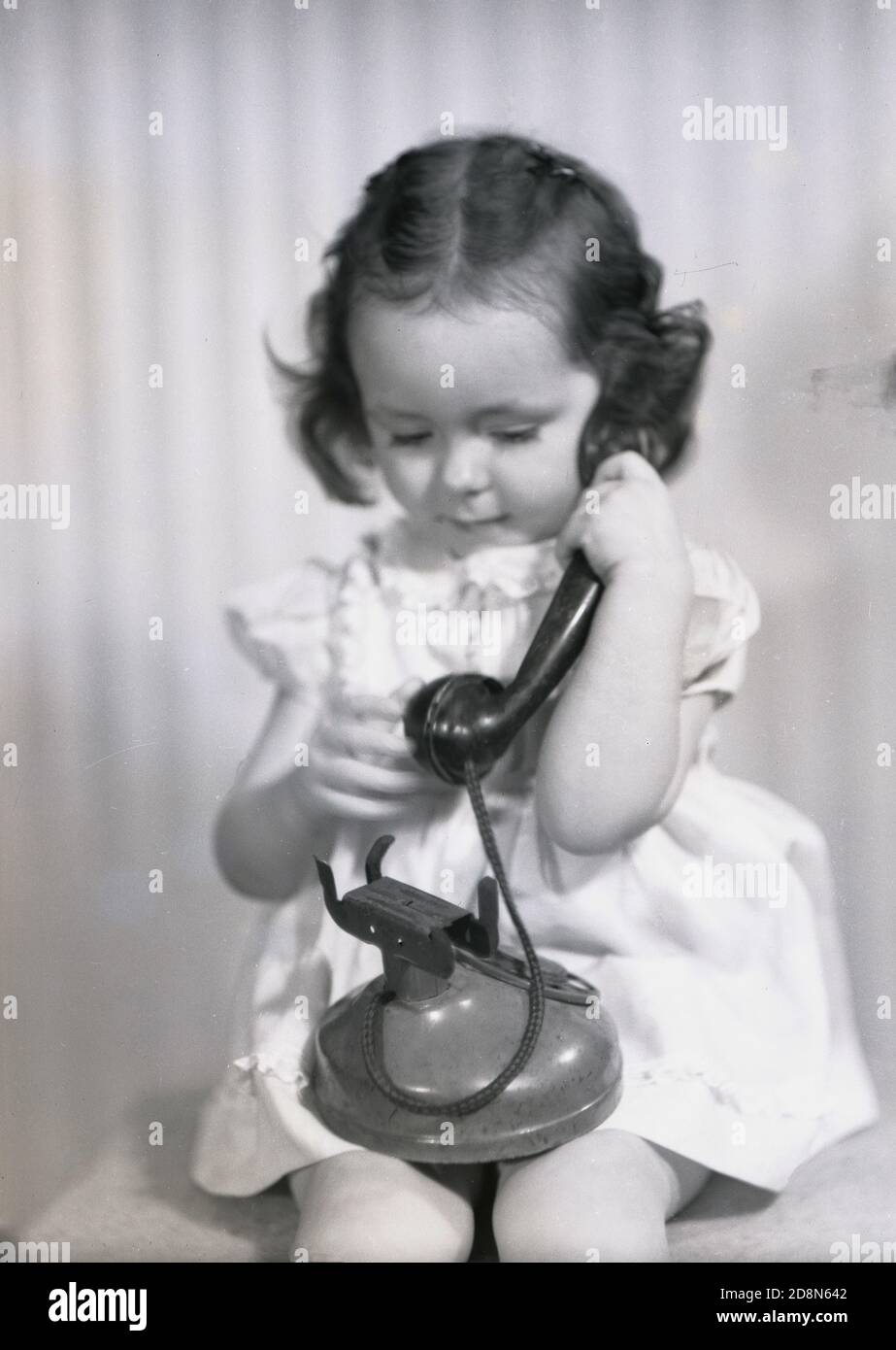 1946, historical, a little girl sitting on a stool playing with a telephone receiver of the era holding it to her ear, as she has seen adults do. Stock Photo