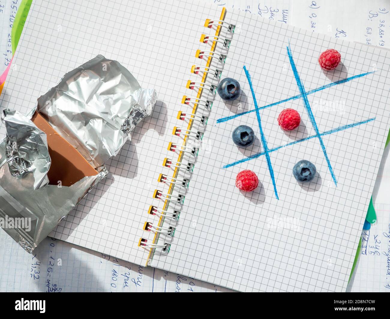 Back to school concept. Tic-tac-toe logic game by raspberries and blueberries on open squared ring-bound notebook near chocolate bar in a silver foil. Stock Photo