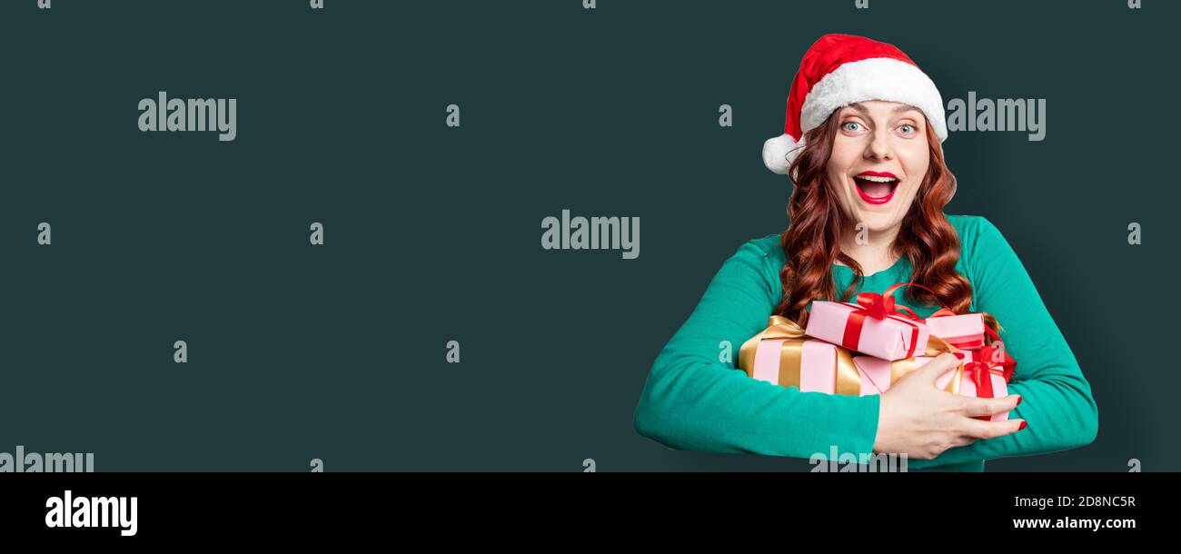 Portrait of pretty santa hat woman holding gift boxes for holiday over green background. Wow celebrating New Year web banner Stock Photo