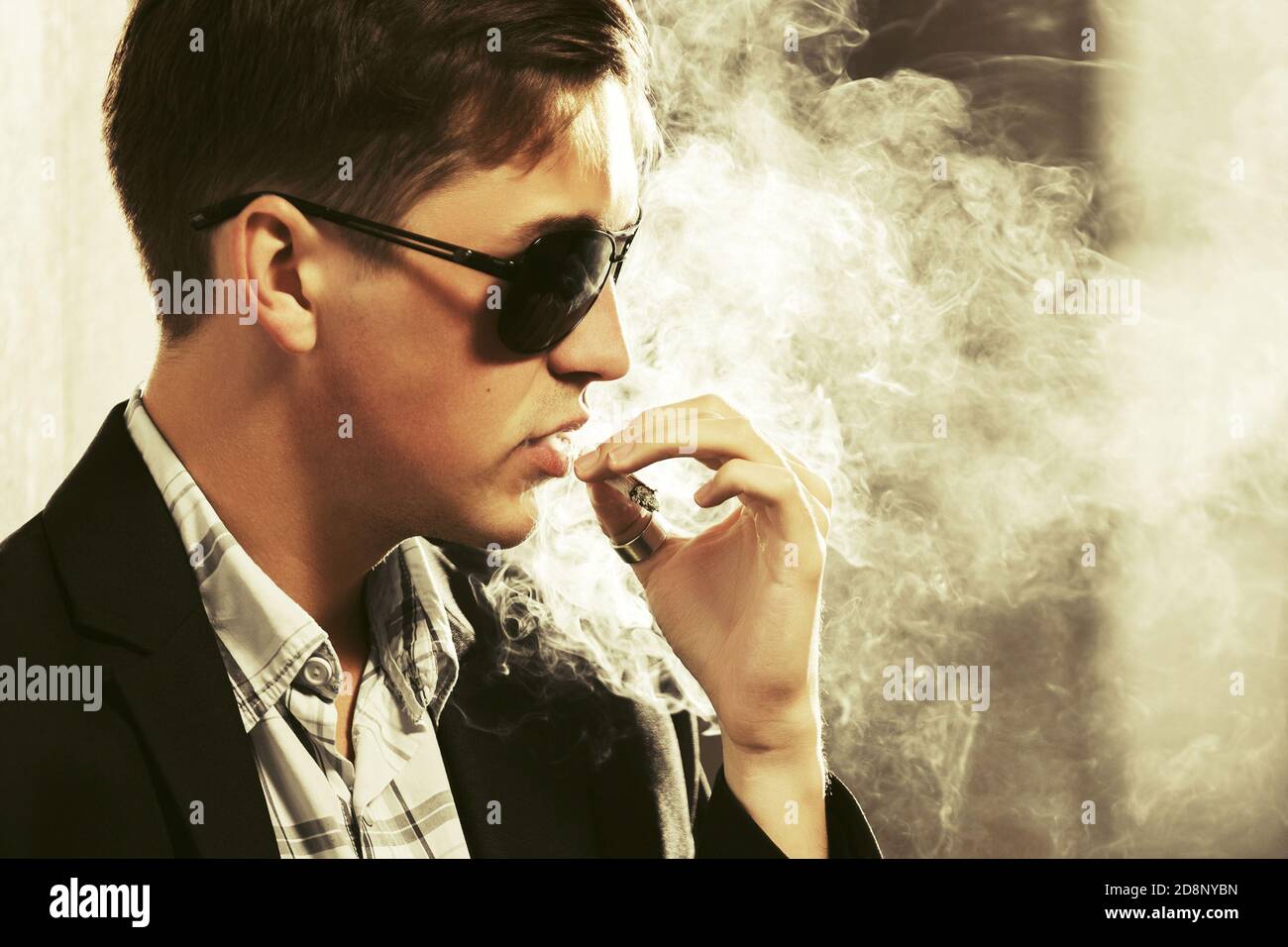 Young man in sunglasses smoking a cigarette on city street  Stylish fashion male model in black suit jacket Stock Photo