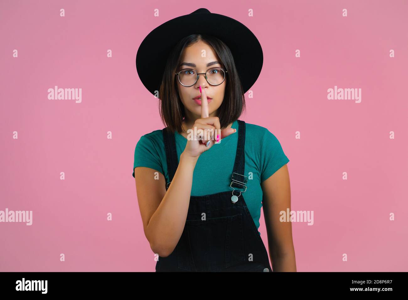 Hipster woman in hat holding finger on her lips over pink background. Gesture of shhh, secret, silence. Close up. Stock Photo