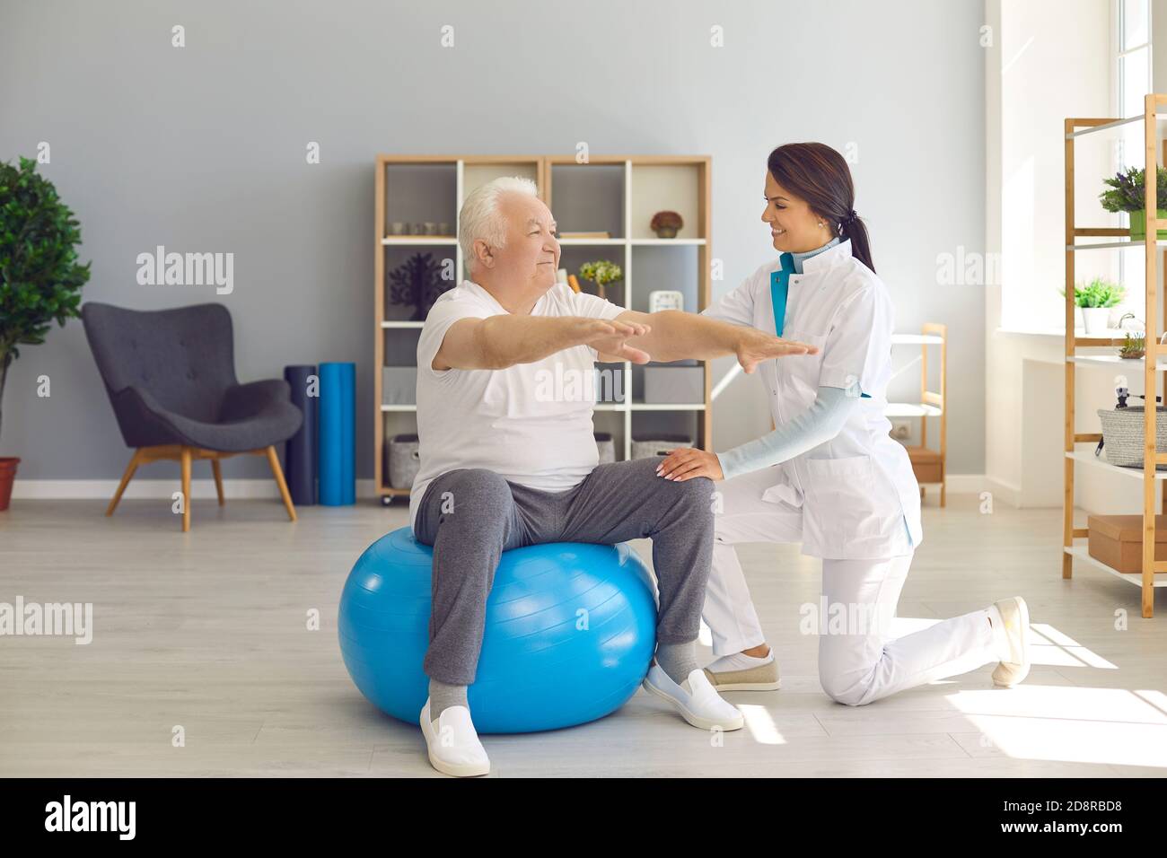 Smiling woman chiropractor correcting old man movements during rehabilitation theapy on fitness ball Stock Photo