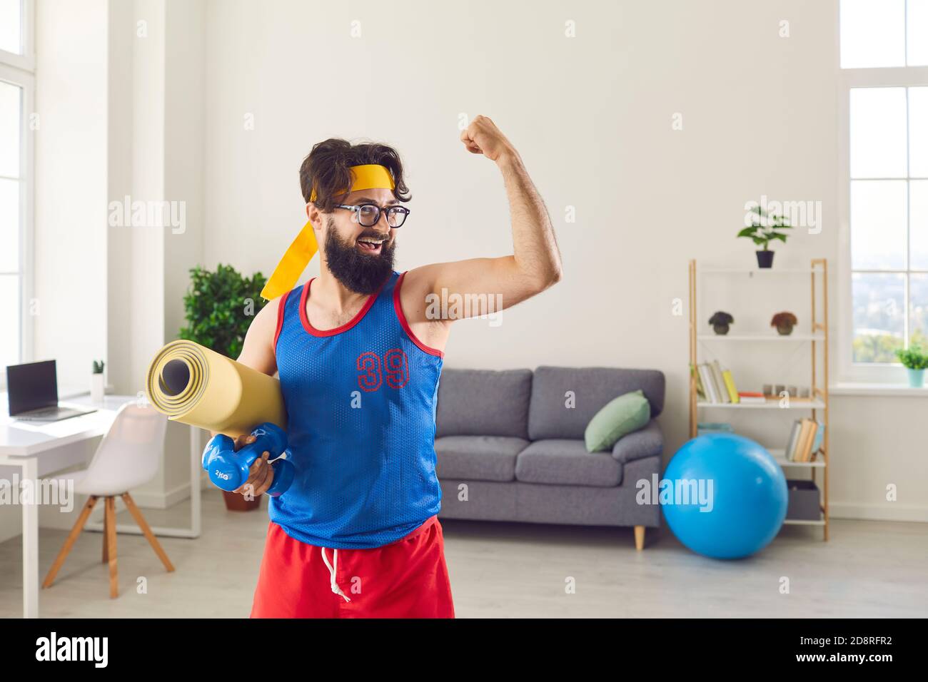 Funny athlete holding gym mat and dumbbells and showing his weak biceps after sport workout at home Stock Photo