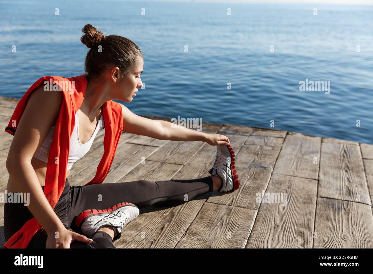 Outdoor shot of attractive fitness woman warming-up before jogging, sitting on pier and stretching legs Stock Photo