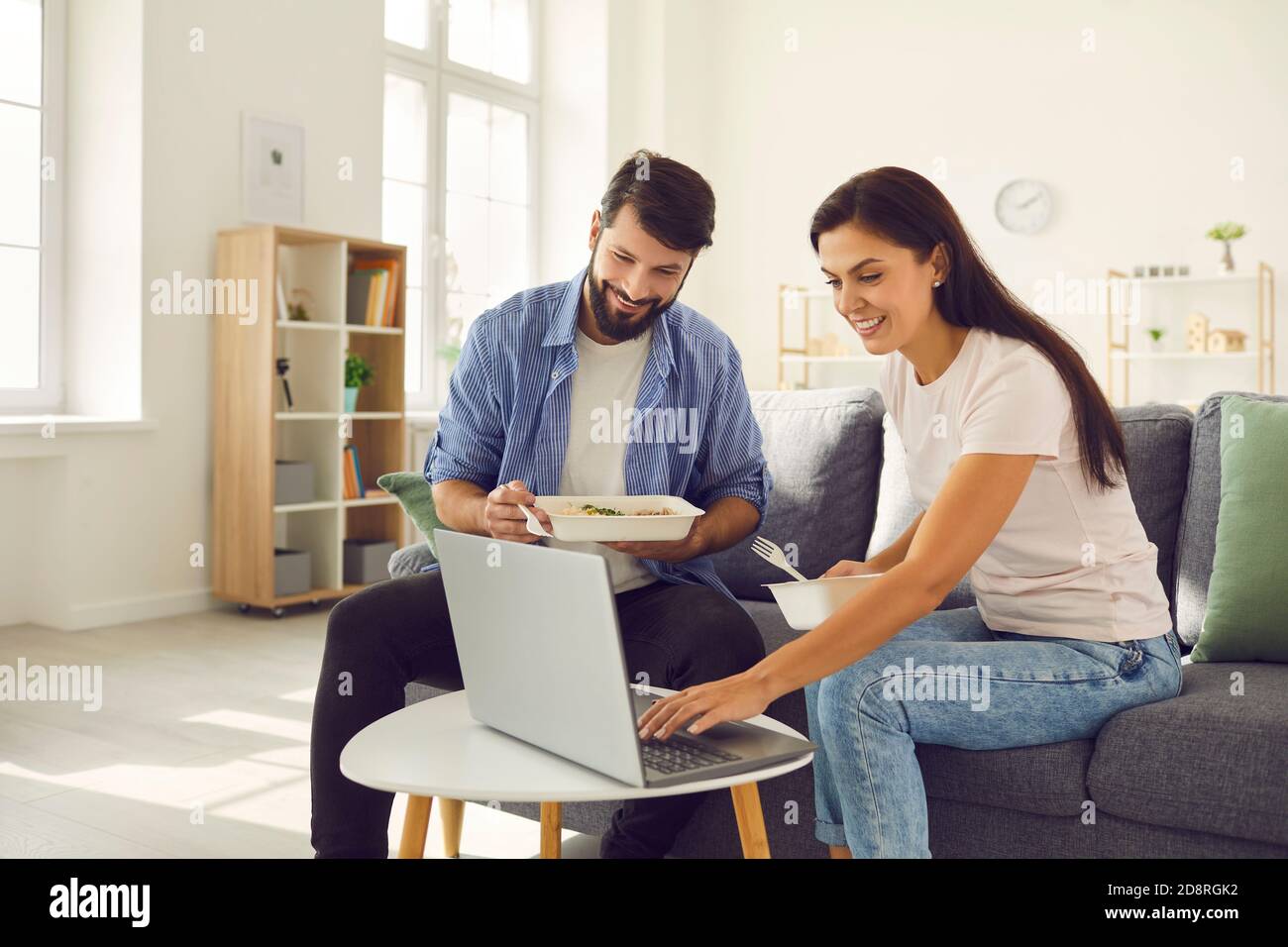 Young man and woman sitting on sofa in the day and eating takeaway food and using laptop. Stock Photo