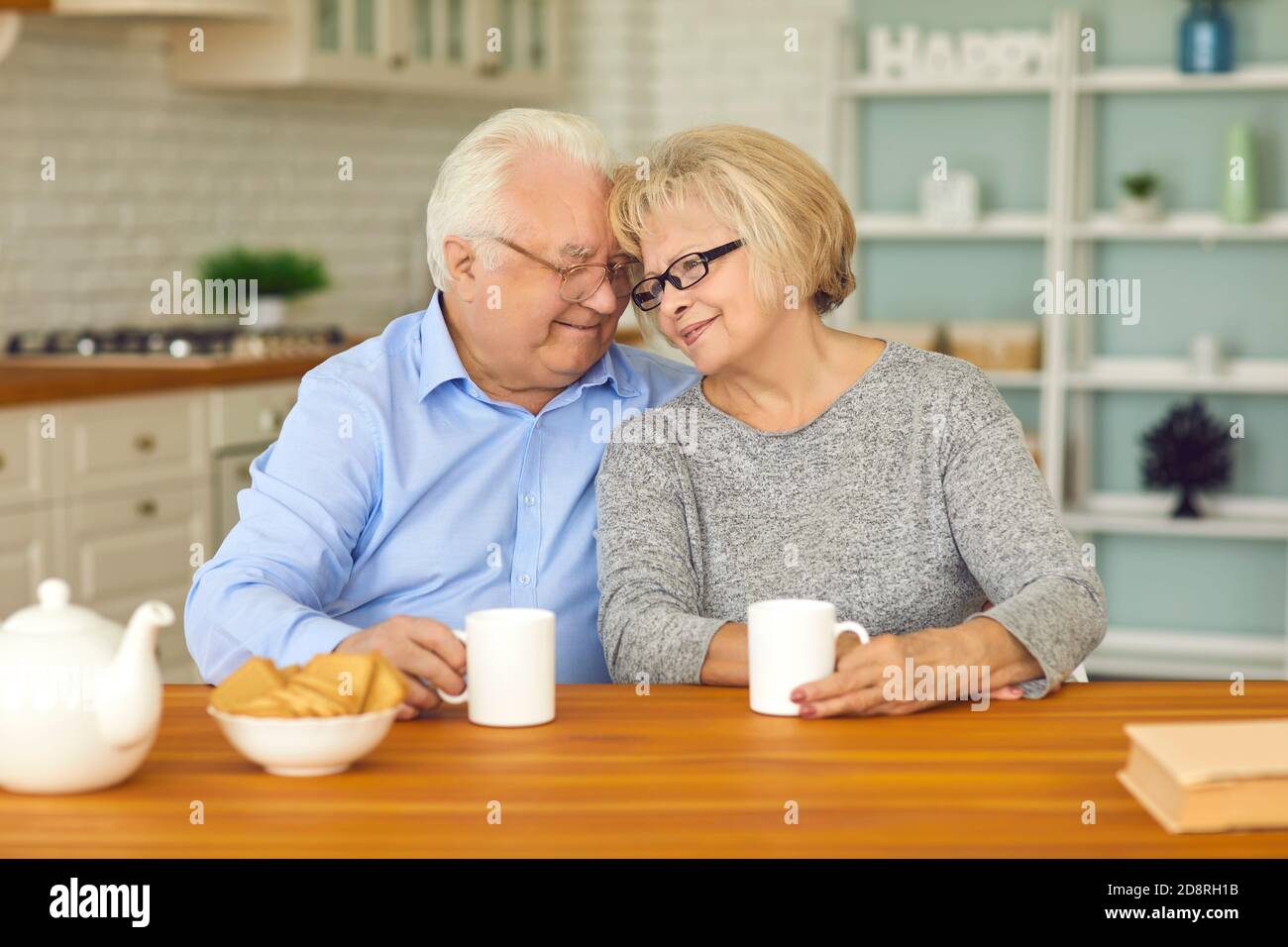 Happy senior couple drinking tea and hugging each other sitting at table in the kitchen Stock Photo