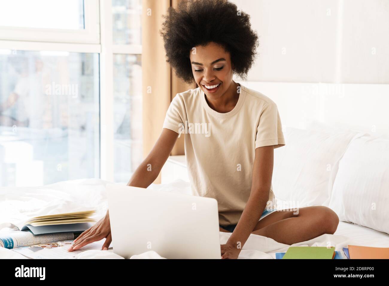Smiling young african girl studying while sitting on bed with laptop ...