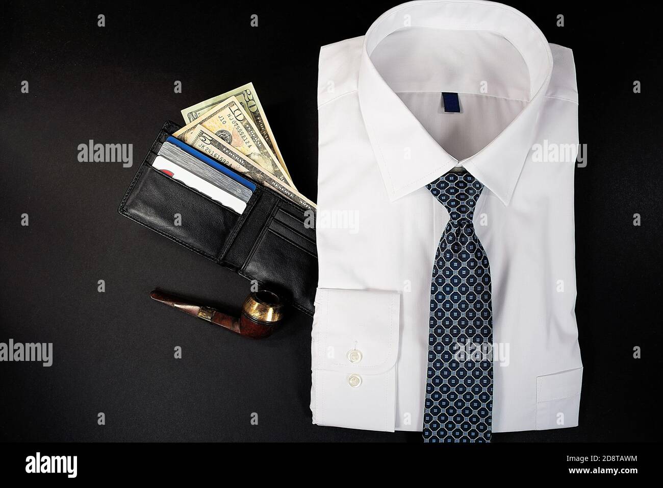folded white dress shirt with necktie and money in black leather wallet and pipe on black background Stock Photo