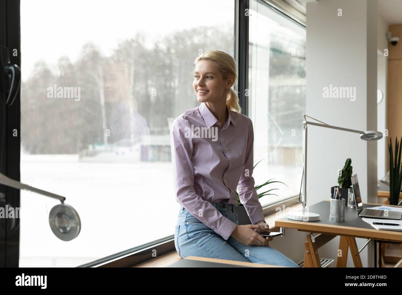 Dreamy young businesswoman sitting on windowsill in office. Stock Photo