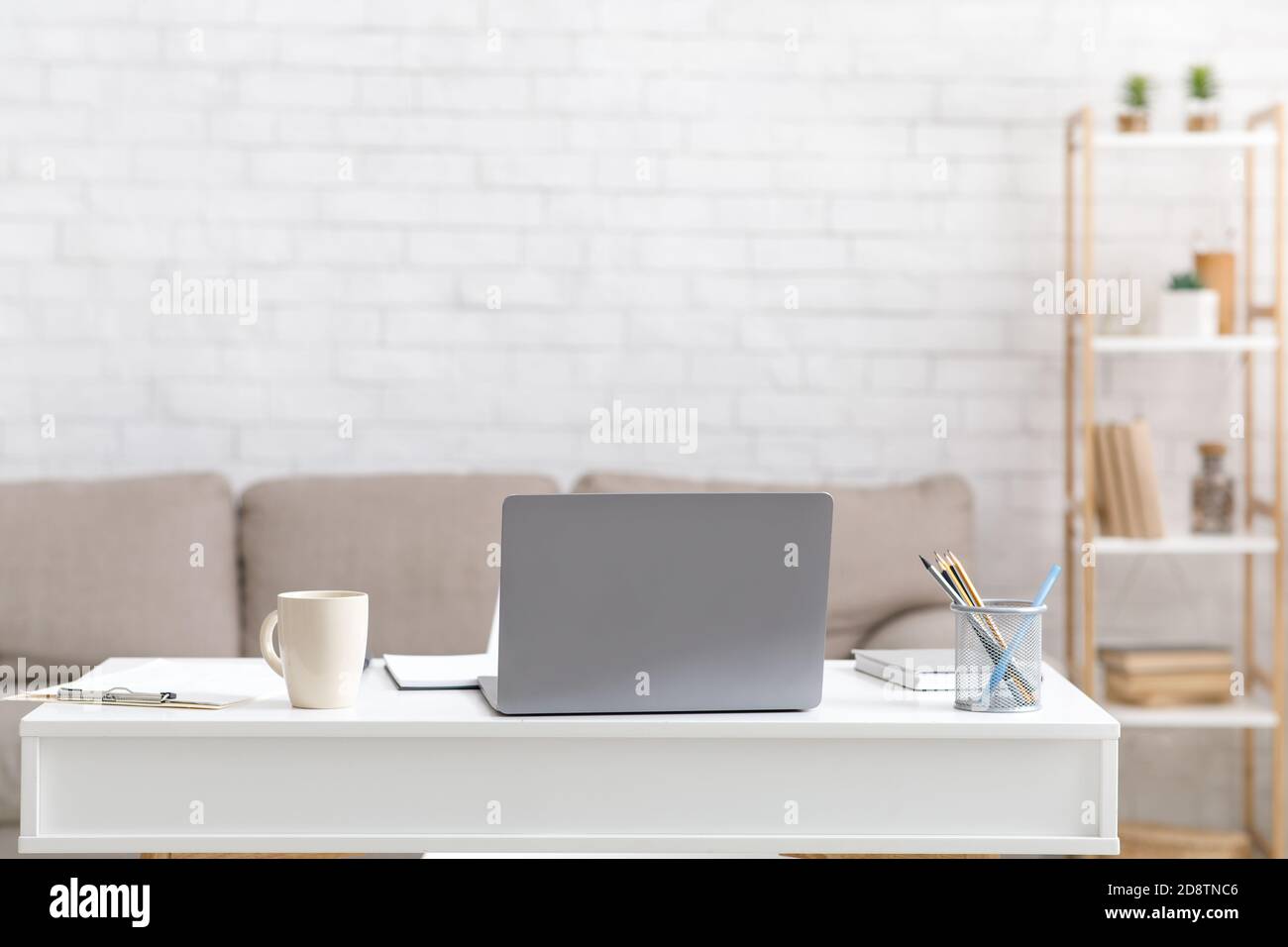 Focus on laptop on table with cup, notepads, pencils, near sofa and white brick wall of house Stock Photo