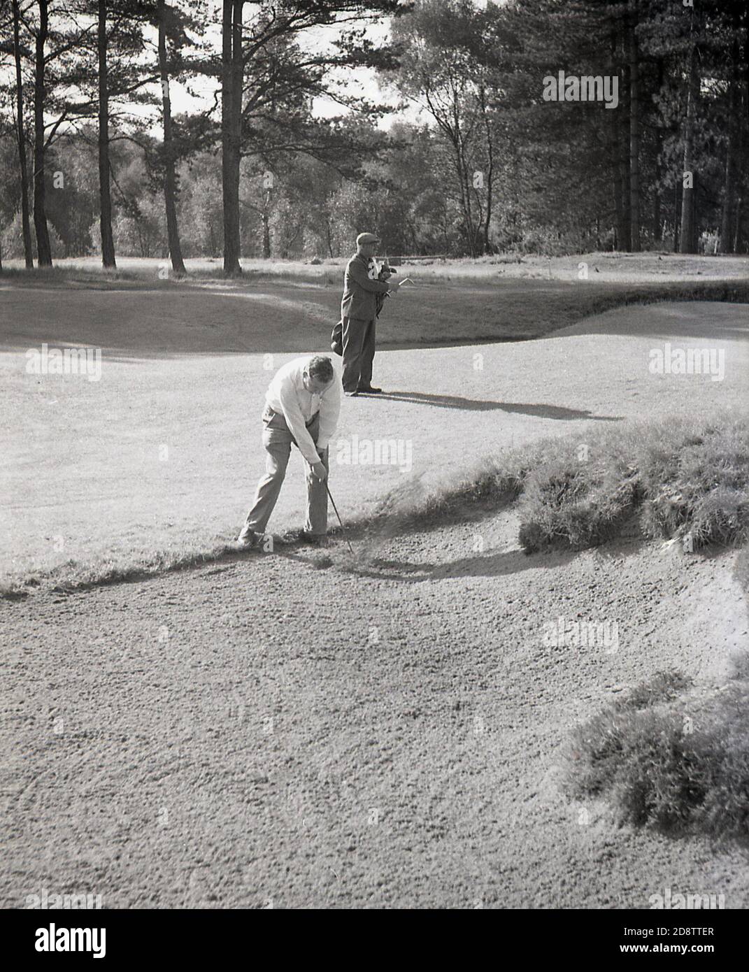 1960s, historical, a male golfer playing out of a fairway or bunker, with caddy standing level with him, carrying his golf clubs, England, UK. Stock Photo