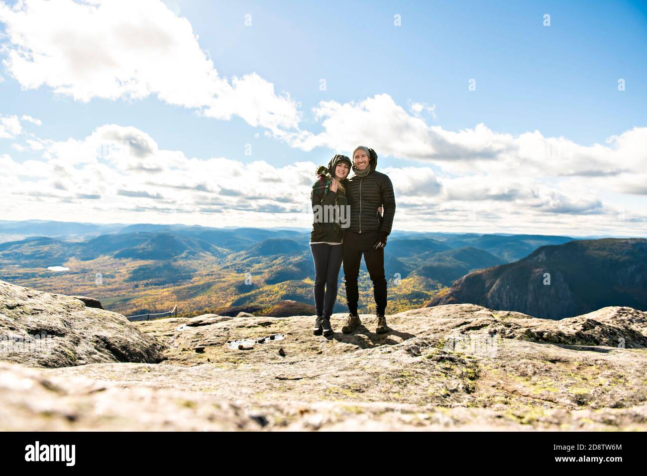 A young tourist couple stand on rocky mountain top enjoying mountain ...