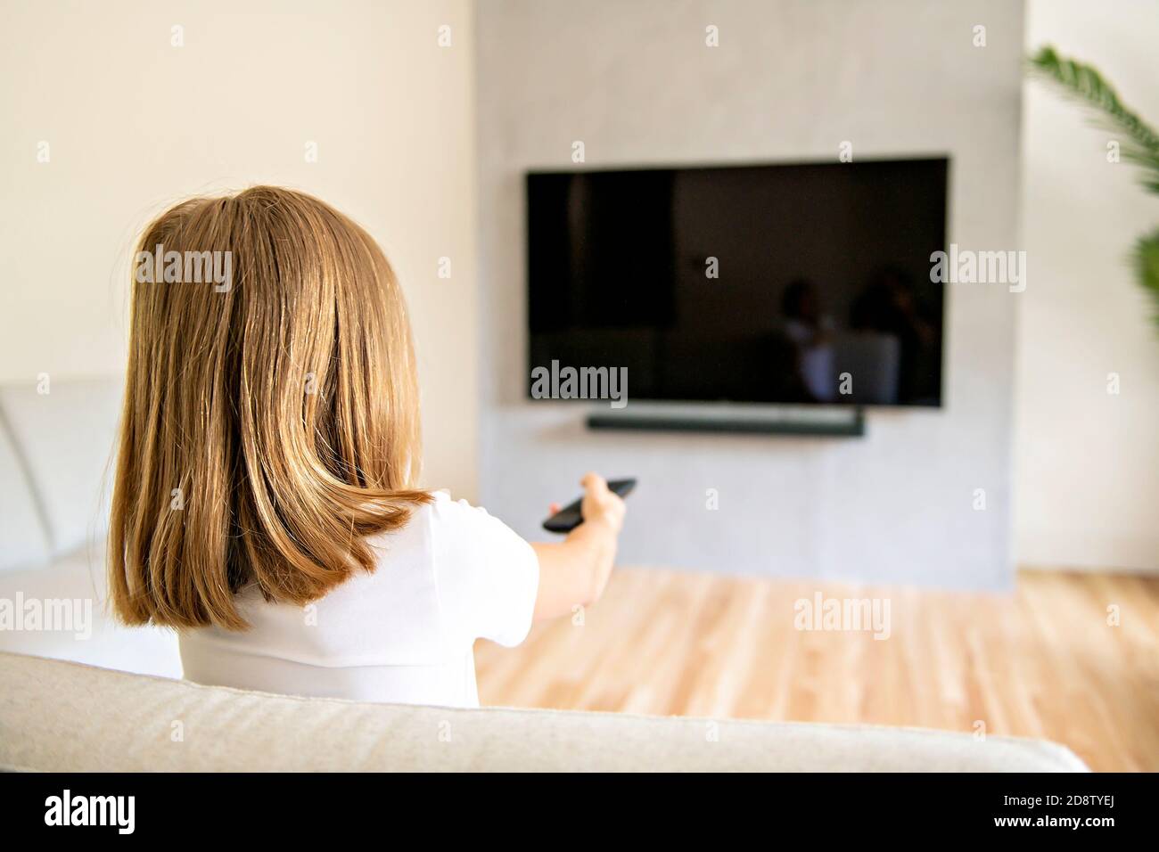 Back view image of cute little blond hair girl sitting on sofa and watching TV. Stock Photo