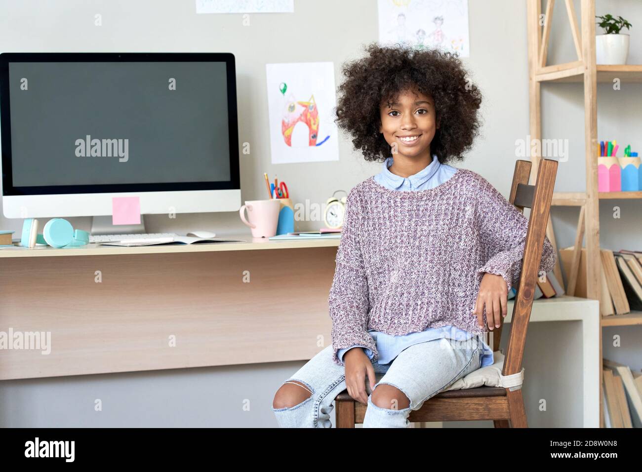 Happy african kid girl looking at camera sitting at home desk with computer. Stock Photo