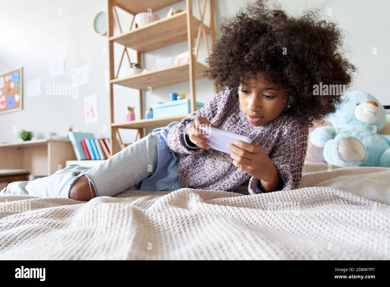 Happy african child girl using mobile apps on smartphone lying on bed at home. Stock Photo