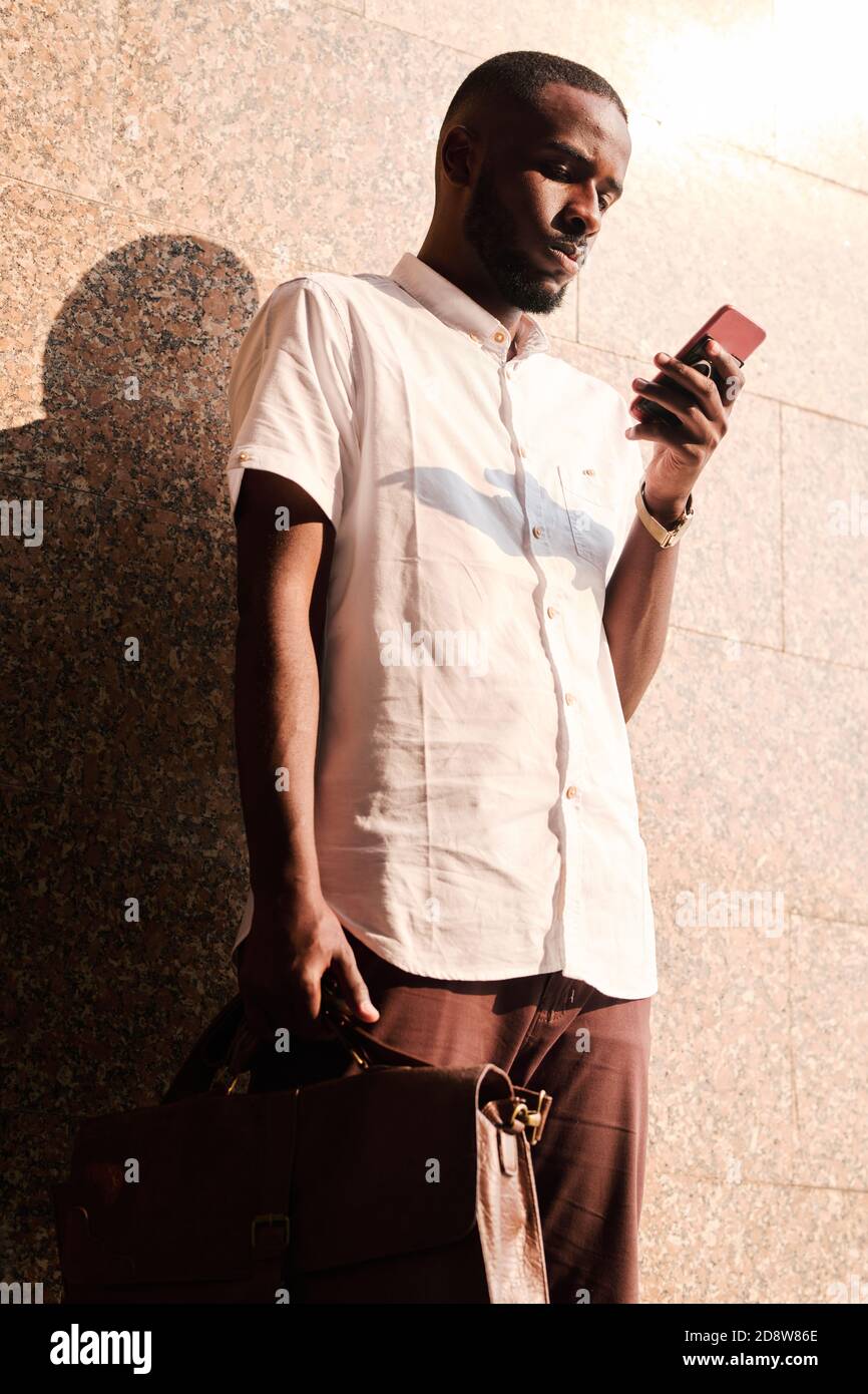 Young serious African businessman in casualwear texting by wall of building Stock Photo