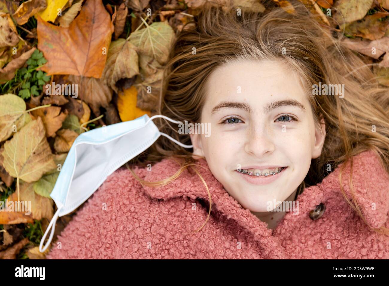 Teenage girl wearing braces smiling and laying in autumn leaves with a face mask besides her. Stock Photo