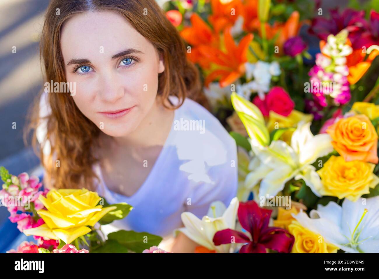 Closeup of beautiful serene young woman with calm expression in a white shirt  surrounded by abundant colorful florist flowers Stock Photo