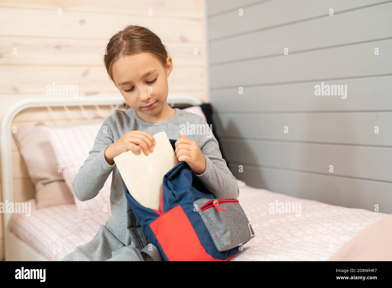 Serious schoolgirl in grey dress putting book into backpack before school Stock Photo