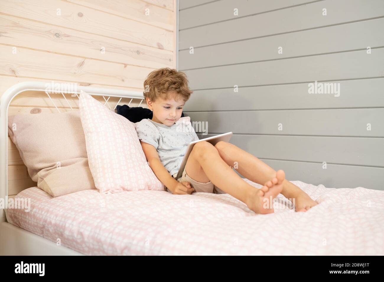 Cute little relaxed schoolboy with touchpad leaning against pillows on bed Stock Photo