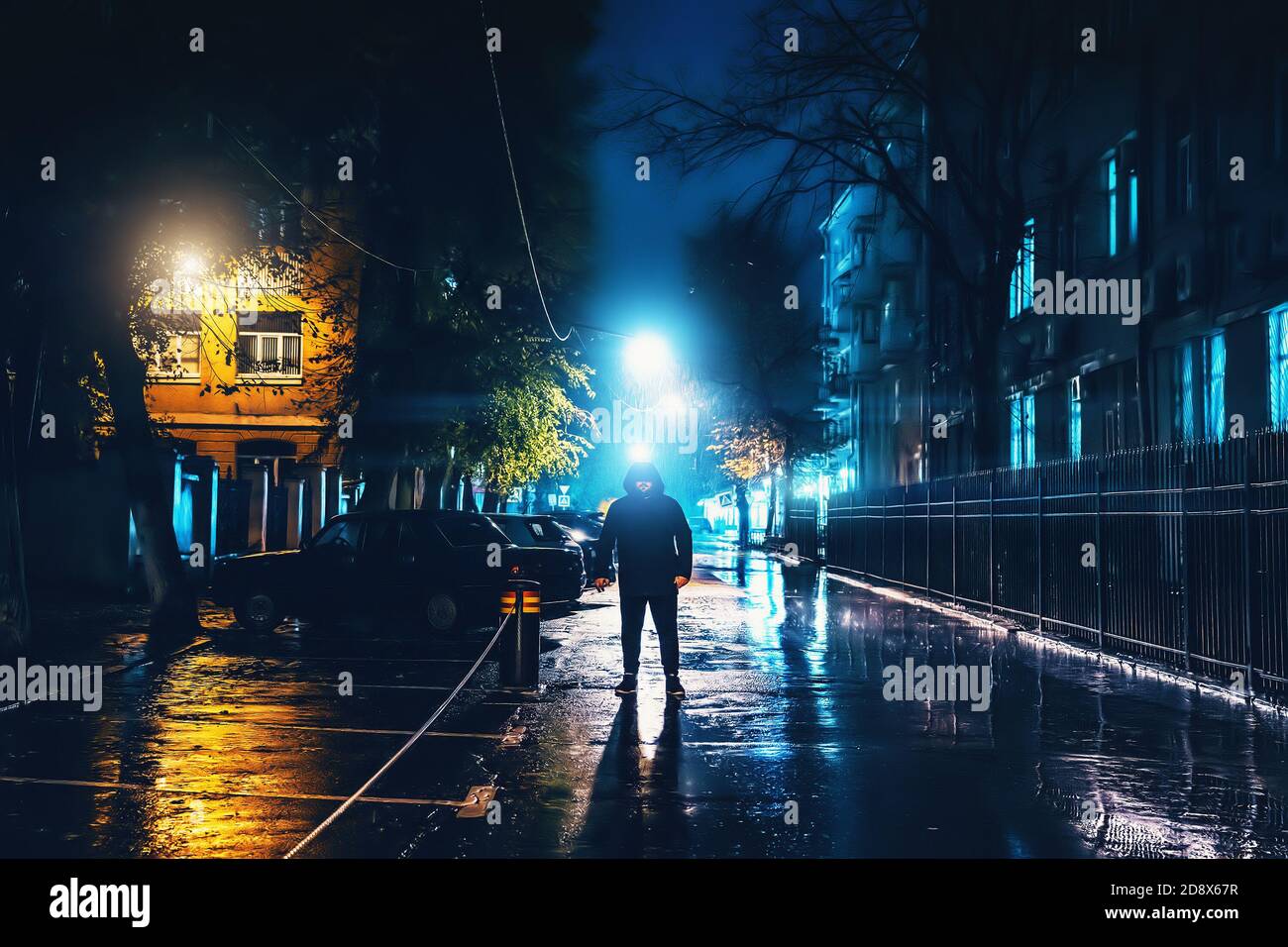 Silhouette of alone stranger in hood at night city street in rain. Creepy killer or stalker, criminal stands in shadow with urban lights reflected in puddles. Thriller horror mysterious atmosphere. Stock Photo