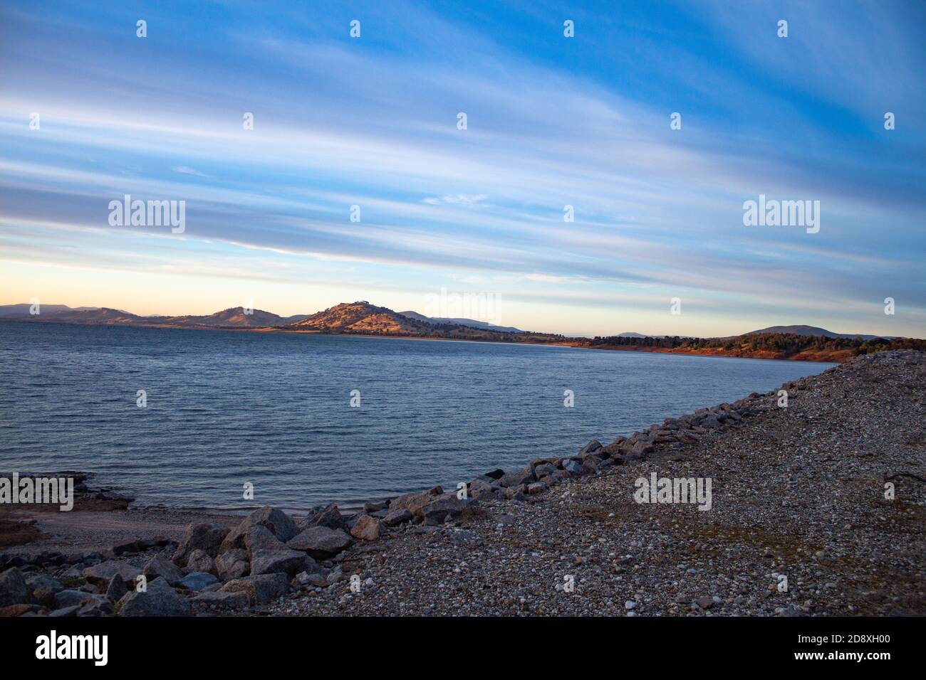 Blue water filled lake dam with rocky foreshore and small hills in background against blue sky Stock Photo