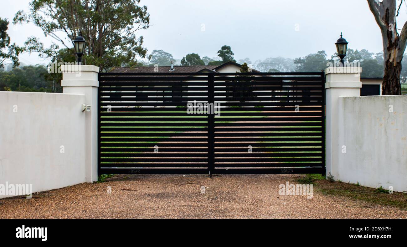 Black modern metal driveway property entrance gates set in concrete fence, lights, trees Stock Photo